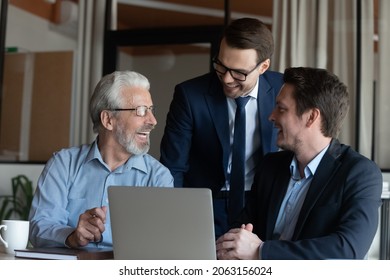 Intergenerational Business Team. Smiling Male Colleagues Of Diverse Age Have Fun By Computer At Workplace. Older And Younger Men Office Employees Gather At Laptop Talk Laugh Joke On Funny Work Moment