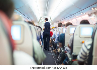 Interior of airplane with passengers on seats and stewardess in uniform walking the aisle. 