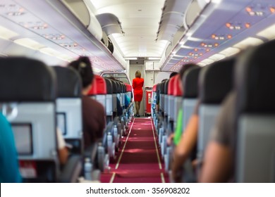 Interior of airplane with passengers on seats and stewardess in red uniform at the aisle. 