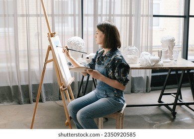 Interior of artist's workshop. Room with huge window. Creative person concept. Easel. Woman paints a picture. Brush and palette in her hands. She is wearing denim overalls and a plaid shirt. - Powered by Shutterstock