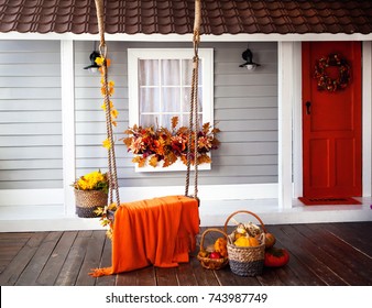 Interior Of An Autumn Patio. Swing Is Adorned With Autumn Leaves And Orange Knitted Plaid. Basket With Pumpkins And Autumn Vegetables. The Window Is Decorated With Autumn Decor. Bord Door.