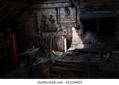 Interior Of Blacksmith Forge With Tools Hanging On The Wall And Anvil And Hammer Ready To Be Used. Furnace Formant, Shaft Of Light From Roof Highlighting The Bricks Of The Wall