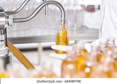 Interior Of Clean Modern Factory Of Creation Cosmetic. A Research Worker Standing Near Manufacturing Facility Holding Glassware With Yellow Substance Creating New Kind Of Cosmetic. Research Concept