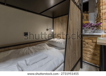 Interior close-up of a hostel bunk bed featuring white linen and a towel. A modern reading lamp illuminates the wood-panelled sleeping space. Next to it, an exposed brick wall and a window with festive city lights create a cozy night atmosphere.