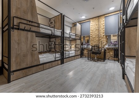 Interior of a contemporary hostel dormitory in loft style at night. Features wooden bunk beds with metal frames, a workspace with a desk and chair, and a brick wall. Bright laminate flooring and cozy lighting create a welcoming space for guests.