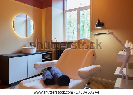 Interior of the cosmetology office with a sink, a round mirror, an electrical facial beauty bed and chair, a trolley cart with skincare products, a led lamp in a beauty salon. A window curtain raised.