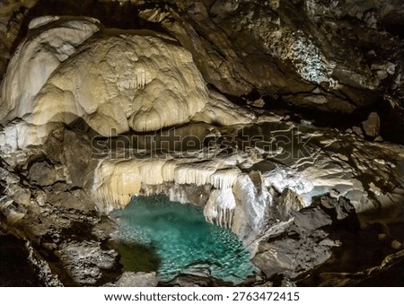 Interior of a deep limestone cave with beautiful stalactites and stalagmites. Underground lake with turquoise water and mineral rock formations illuminated by artificial light. New Athos, Abkhazia.