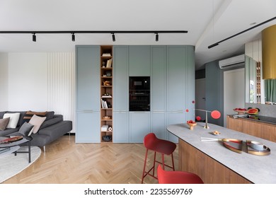 Interior design of colorful open space with built-in oven, bookcase, modern red hockers, wooden kitchen island, gray sofa, pillows, panels floor and personal accessories. Home decor. Template.