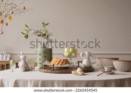 Interior design of easter dining room interior with round table,  colorful eggs, easter cake, easter bunny sculptures, vase with flowers, beige bowl and personal accessories. Home decor. Template.