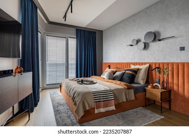 Interior design of elegant bedroom with big orange bed, beige and grey bedclothes, blue curtain, rug, modern lamp, night stand, vase with dried flowers and personal accessories. Home decor. Template. - Shutterstock ID 2235207887