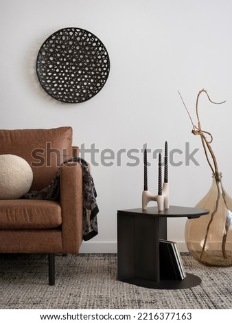 Interior design of harmonized living room with brown sofa, coffee table, glass vase with dried flowers, round pillow, blanket, candlestick  carpet and personal accessories. Creative home decor.