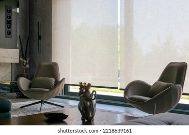 Interior design with roller blinds in the background. Automatic solar shades of large sizes on the window. Fabric with linen texture. In front of a large window is a chair on a carpet. 