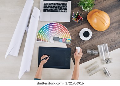 Interior Designer Working On Graphic Tablet At Desk With Color Palette, Blueprints And Cup Of Coffee, View From Above