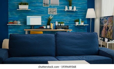 Interior Of Elegant Bright Living Room With Work Place And Big Television In Background. Confortable Couch In Empty Modern Apartment With Blue Furniture And Walls, Beautiful Decorated And No People In