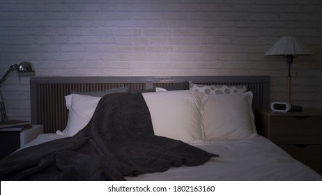 Interior Of Empty Bedroom At Night, With Unmade Bed And Tv Left On At Modern Living Space With White Bedding And Grey Blanket 