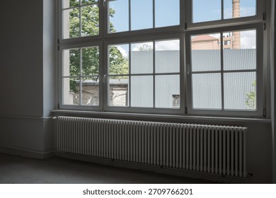 Interior of an empty room featuring a large multi-pane window above a long white radiator, offering a view of green trees and an industrial building with a brick chimney.