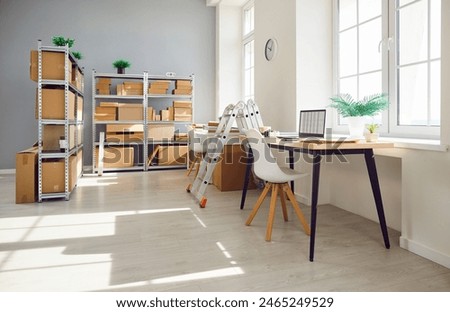 Interior of empty work space with office desk in warehouse with shelves filled cardboard boxes of retail products. Business workplace used for shipping merchandise and stock inventory.