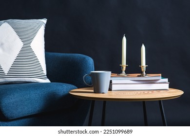 interior and home decor concept - close up of blue chair with pillow, candles, books and mug on coffee table in dark room