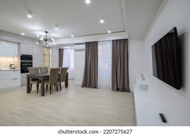 Interior Of A Huge White Luxury Kitchen In A Studio Apartment, Tv Set, Lots Of Copy Space, Large Windows With Curtains And A Dining Table With Chairs