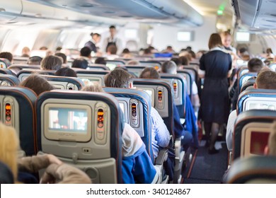 Interior of large passengers airplane with people on seats and stewardess in uniform walking the aisle. 