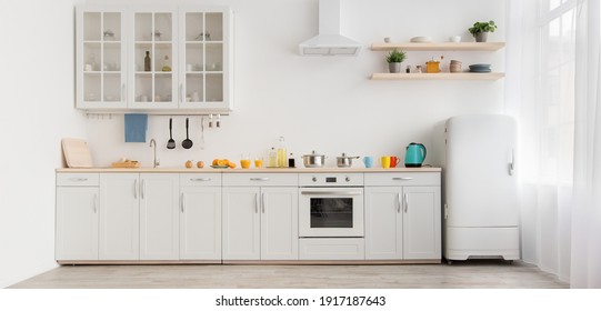 Interior of modern comfortable kitchen. Multicolored cups and teapot, orange juice in glass and utensils on white furniture, refrigerator, flowers in pots on shelves, light wall in daylight, panorama