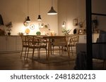 Interior of modern kitchen with glowing lamps, white counters, chairs and dining table at evening