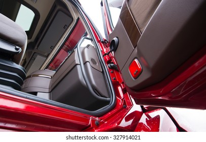 The Interior Of Modern Luxury Red Semi Truck Made In Shades Of Brown Plastic. Through Open Door With Wood Paneling Truck Visible Part Of Seat With Shock Absorbs And Dashboard Console With Air Vents.