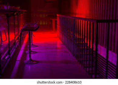 Interior Of A Nightclub With Colorful Red And Purple Lighting And A Line Of Stylish Bar Stool Along A Reflective Bar Counter With A Railing Behind