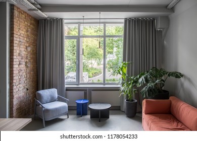 Interior In An Office In A Loft Style With Gray And Brick Walls. There Is A Peach Sofa, Green Plants In Big Pots, Small Round Tables, Light Armchair, Window With Curtains, Hanging Lamps. Horizontal.