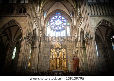 Interior of old catholic church with golden altar in centre, large stained-glass window over it, huge arches and high columns made of grey stone. Religion and faith. Holy places and architecture.