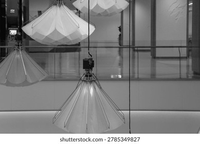 An interior perspective photograph capturing multiple modern geometric-patterned pendant lights hanging from the ceiling of a luxury shopping mall. On the left, a large, intricate wooden bookshelf spans floor-to-ceiling, filled with various books and decorative items. In the foreground, a sleek wooden counter is equipped with two desktop monitors next to a private study cubicle enclosed by glass partitions with blurred frosted details, under a clean linear ceiling lighting layout. The natural light from the distant background window illuminates the wood textures and the polished floor.
