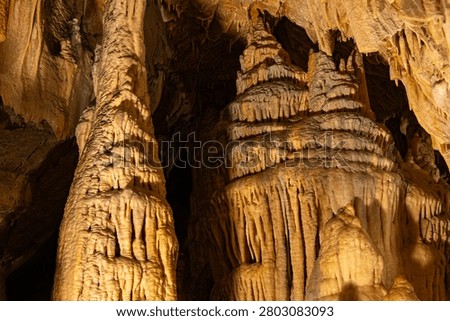 An interior photograph capturing the colossal limestone formations inside the Obir Dripstone Caves (Obir Tropfsteinhöhlen), featuring a large floor-to-ceiling pillar column on the left and a layered stalagmite structure illuminated by dramatic cave lighting