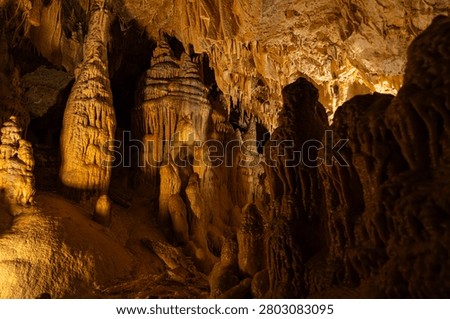 An interior photograph capturing the colossal limestone formations inside the Obir Dripstone Caves (Obir Tropfsteinhöhlen), featuring a large floor-to-ceiling pillar column on the left and a layered stalagmite structure illuminated by dramatic cave lighting
