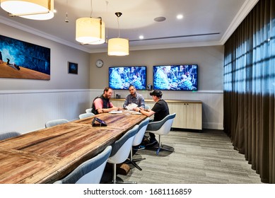 Interior Photography Of A Contemporary Board Room With A Rustic Timber Meeting Table, Swivel Chairs, Pendant Lighting & Timber Cabinetry, Wall Mounted Screens And Curtains, Staff Holding A Meeting