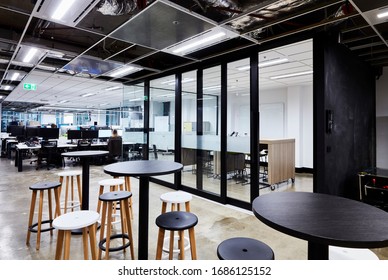 Interior Photography Of A Contemporary Design Corporate Office Break Out Area With Tables & Stools, A Meeting Room And Open Plan Office In The Background With Staff At Their Desks