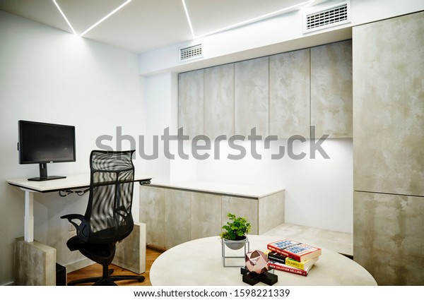Interior photography of a corporate fit out\
corner office with desk and chair, concrete finish cabinetry, round\
table meeting area with books, plant and decorative objects and\
ceiling strip lighting