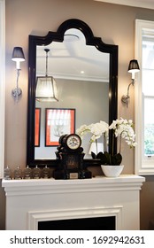 Interior Photography Detail Of A Hamptons Style Lounge Room With A Black And Taupe Decor, A Black Lacquered Mirror Hangs Over A Fireplace With A Clock And Phalaenopsis Orchid