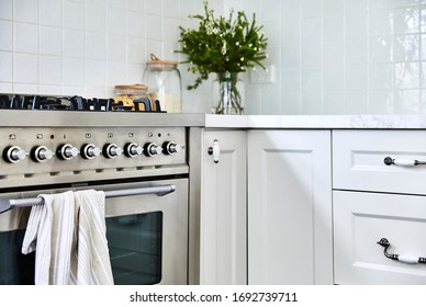 Interior Photography Detail Of White Hamptons Style Kitchen Cabinets With A Marble Bench Top, A Stainless Steel Oven And Stove And A Floral Arrangement In Background