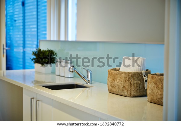 Interior photography detail of a white laundry with\
a caesar stone bench top, a glass splash back with a sink, baskets,\
laundry powder and a\
plant