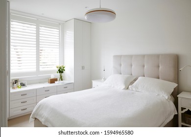 Interior photography of fresh and light bedroom with white bed, upholstered bed head, white built in cupboards and plantation shutters with vase of roses and decorative objects