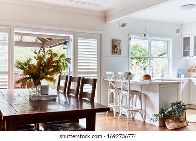 Interior Photography Of A Fresh White Hamptons Style Kitchen With Breakfast Bar, Cross Back Bar Stools, Polished Floor Boards And A Rustic Timber Dining Room Setting To The Left