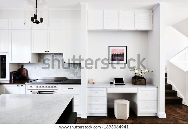 Interior photography of a fresh white\
kitchen and home office area with stairs on the\
right