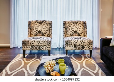 Interior Photography Of Hamptons Style Lounge Room With Printed Upholstered Arm Chairs, Coffee Table With Tea Setting, Rug And Dark Fabric Sofa On Far Right