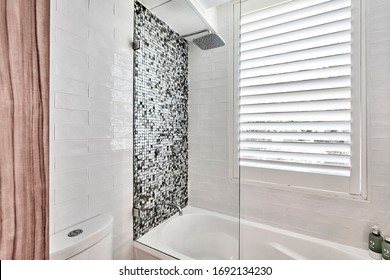 Interior photography of a modern bathroom with a glass mosaic tiled shower over a white bath with a large shower head, white subway tiles, a window with plantation shutters and toiletries