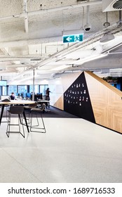 Interior Photography Of A Modern Corporate Office With A Tall Meeting Table And Stools, A Graphic Notice Board Wall And Polished Concrete Floors With Staff In The Background