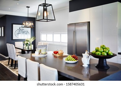 Interior Photography Of Modern Hamptons Style Kitchen With Stone Bench Top Breakfast Bar, Glass Pendant Lighting, Stools, Decorative Objects, Stainless Steel Fridge And Dining Room In Background