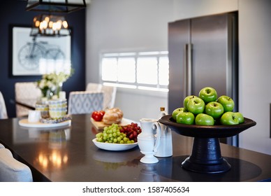 Interior Photography Of Modern Hamptons Style Kitchen With Stone Bench Top Breakfast Bar, Glass Pendant Lighting, Food, Decorative Objects, Stainless Steel Fridge And Dining Room In Background