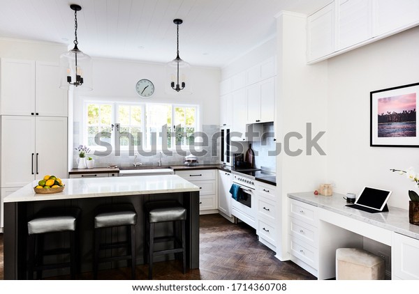 Interior photography of a modern Hamptons style\
kitchen with a marble bench top, pendant lighting and a home office\
media nook area to the\
right