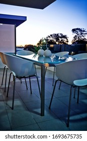 Interior Photography Of A Modern Inner City Apartment Balcony Area At Dusk With Outdoor Table And Chairs And Trees In Distant Background