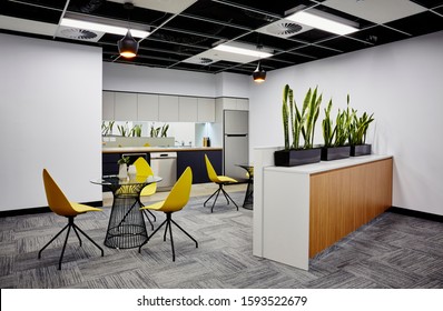 Interior Photography Of Modern Office Break Out Area With Kitchen, Pendant Lighting, Metal And Glass Topped Tables, Yellow Moulded Plastic Chairs, Planter Boxes With Sansevieria Plants